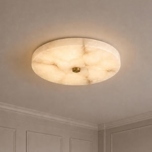 Op de afbeelding: Een ronde plafondlamp voor vlakke montage met een doorschijnende, crèmekleurige stenen kap met natuurlijke aders. Het midden van het armatuur is bevestigd met een kleine, ronde, messingkleurige metalen dop.