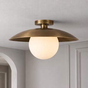 May include: A semi-flush mount ceiling light fixture featuring a wide, shallow, brushed brass metal dome shade. A large, spherical, frosted white glass globe is centered beneath the metal shade, providing a soft, diffused light source.
