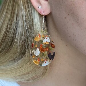 May include: A pair of teardrop-shaped dangle earrings with a clear background and a colorful pattern of chickens and chicks.