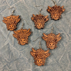 May include: Six wooden Highland cow earrings with silver-coloured ear wires. The earrings feature detailed laser-cut designs of the cow's face, with some adorned with floral crowns. The earrings are displayed on a light blue textured surface.