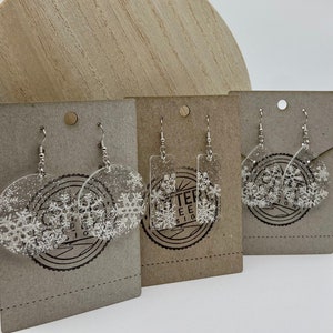 May include: Three pairs of snowflake earrings on brown card stock. The earrings are made of clear resin with silver glitter and white snowflake designs. The earrings are on silver hooks. The card stock has the text "Glitter Tree Designs" printed on it.