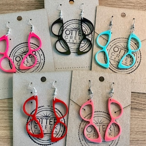 May include: Four pairs of earrings shaped like glasses. The earrings are in four different colors: black, pink, red, and turquoise. Each pair of earrings has the text "Otter Creek Design" on the back.