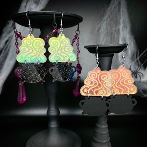 May include: Two pairs of iridescent Halloween earrings. The first pair features a green cauldron with swirling smoke and a black cauldron charm. The second pair features an orange cauldron with swirling smoke and a black cauldron charm.