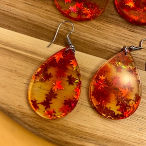 Peut inclure: Une paire de boucles d'oreilles en forme de goutte d'eau avec une base en résine transparente et des paillettes orange et rouges incrustées à l'intérieur. Les paillettes ont la forme de feuilles.