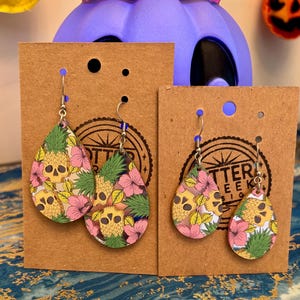 May include: Teardrop earrings with a skull and pineapple design. The earrings feature a yellow skull, pink flowers, and green pineapple leaves on a light pink background. The earrings are displayed on brown card.