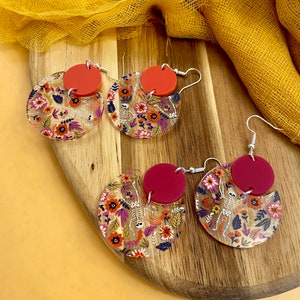 May include: A pair of earrings with a floral and skeleton design. The earrings are made of clear acrylic with a red circle in the center. The floral design is in shades of orange, pink, and purple.