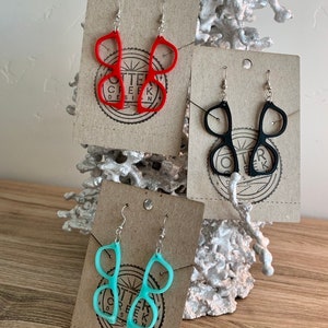 May include: Three pairs of earrings shaped like glasses. The earrings are displayed on brown cards with the text "Otter Creek Design". The earrings are red, black, and light blue.