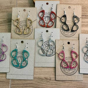 May include: Assortment of glitter eyeglasses earrings in various colors, including gold, silver, pink, teal, and black. Each pair is displayed on a card with the text "Otter Creek Design".