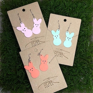 May include: Three sets of earrings featuring a pink, light blue, and orange bunny shaped charm. Each set is displayed on a card with the text "Otter Creek Creations".