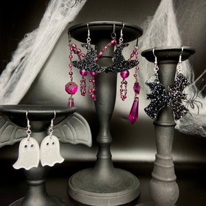 May include: Three pairs of Halloween-themed earrings displayed on black stands. The earrings are made of black glitter and feature a witch's hat, a bat, and a ghost.