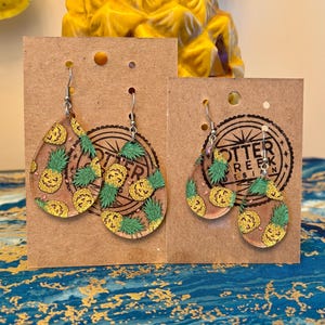 May include: Two pairs of teardrop-shaped earrings with a pineapple design. The earrings are clear with a gold glitter pineapple pattern and green leaves. They hang from silver-colored hooks and are displayed on brown cardboard.