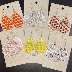 May include: Six pairs of dangle earrings with different sports ball patterns. The earrings are made of clear acrylic with a silver hook. The patterns include basketball, volleyball, football, baseball, softball, and soccer.