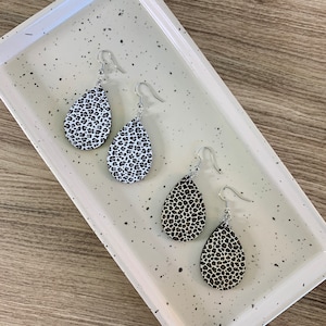May include: Three pairs of dangle earrings with a teardrop shape. The earrings are made of white and black leopard print material. The earrings are on a white tray with black speckles.