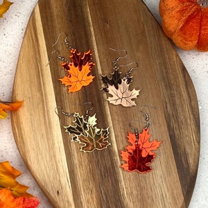 May include: Three pairs of dangle earrings featuring a layered leaf design. The leaves are made of mirrored acrylic in shades of orange, gold, and copper.