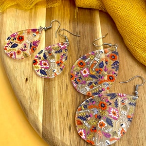 May include: Clear, teardrop-shaped earrings with a floral and skeleton design. The earrings feature a mix of orange, pink, and blue flowers with white skeleton figures. The earrings are displayed on a wooden surface.