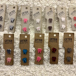 May include: A set of 10 pairs of tooth-shaped earrings with various patterns, including polka dots, stripes, and floral designs. The earrings are made of wood and have a natural finish. The earrings are displayed on small cards with the text "Otter Creek Design".