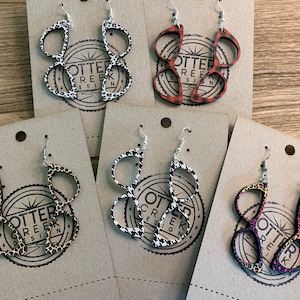 May include: Four pairs of wooden earrings with different patterns. The earrings are shaped like a pair of glasses and are layered on top of each other. The patterns include leopard print, black and white houndstooth, red and black plaid, and a multi-colored pattern. The earrings are labeled "Otter Creek Design".