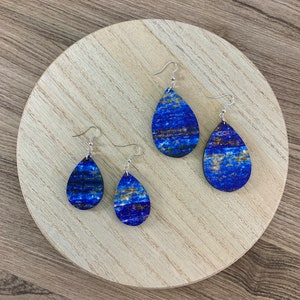 May include: Four pairs of teardrop-shaped earrings with a blue and gold marbled design. The earrings are displayed on a light wooden round tray. The earrings have silver-colored hooks.
