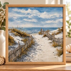 May include: A square cross-stitch embroidery in a light wood frame depicting a sandy beach path leading to the ocean. The scene features white sand dunes, golden beach grass, and a weathered wooden fence under a blue sky with white clouds.