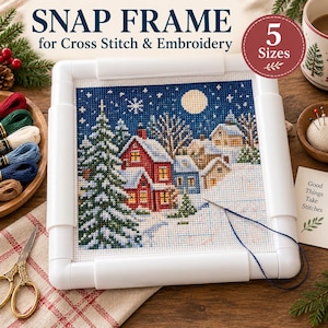 May include: A white plastic snap frame for cross stitch and embroidery holding a fabric piece with a winter landscape design featuring houses, a large pine tree, and a full moon. The text SNAP FRAME for Cross Stitch & Embroidery is visible.