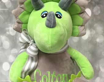 May include: A green and gray plush dinosaur toy with a white and gray striped bow. The dinosaur is sitting with its legs crossed and has the name "Colton" embroidered on its chest. The text "Monogram Perfect" is visible in the bottom right corner of the image.