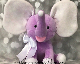 May include: A plush purple elephant toy with large floppy ears, a white bow, and a pink tongue sticking out. The elephant is sitting on a gray background.