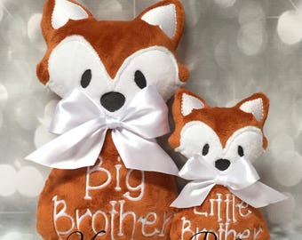 May include: Two plush fox toys, one larger than the other. The larger fox is brown with white accents and a white bow. It is embroidered with the text "Big Brother". The smaller fox is also brown with white accents and a white bow. It is embroidered with the text "Little Brother". Both foxes are sitting on a gray background.