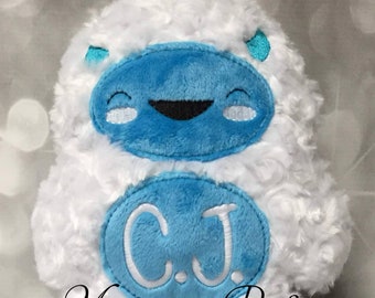 May include: White plush yeti with blue eyes and a big smile. The yeti has a blue oval on its chest with the letters "C.J." embroidered in white.