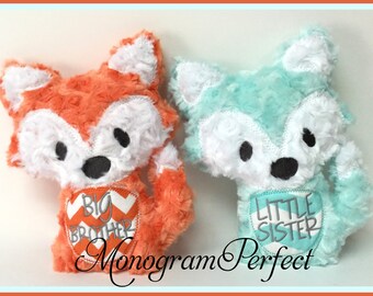 May include: Two plush fox toys, one orange and one light blue, with embroidered white faces and black eyes. The orange fox has a chevron heart with the text "Big Brother" and the blue fox has a chevron heart with the text "Little Sister".