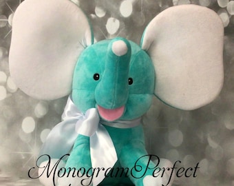 May include: A turquoise plush elephant with white ears and a white bow. The elephant has a pink tongue sticking out and black eyes. The text "Monogram Perfect" is visible in the bottom right corner of the image.