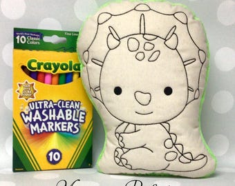 May include: A plush dinosaur-shaped pillow with black outline details and a green border, next to a box of Crayola Ultra-Clean Washable Markers with 10 classic colors. The pillow is designed for coloring.