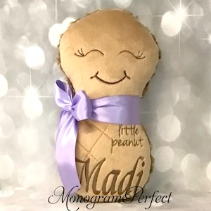 Puede incluir: Un juguete de peluche color canela con forma de cacahuete, con ojos cerrados y boca sonriente bordados. El juguete tiene una cinta de sat&eacute;n lavanda atada alrededor del centro, con el texto "little peanut" y el nombre "Madi" bordados al frente.