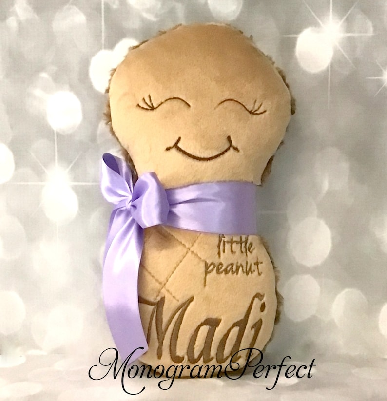 Puede incluir: Un juguete de peluche color canela con forma de cacahuete, con ojos cerrados y boca sonriente bordados. El juguete tiene una cinta de sat&eacute;n lavanda atada alrededor del centro, con el texto "little peanut" y el nombre "Madi" bordados al frente.