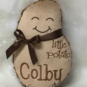 May include: A brown plush potato shaped cushion with a smiling face embroidered on it. The cushion has a brown satin ribbon tied around it and the words "little potato Colby" are embroidered on the front.