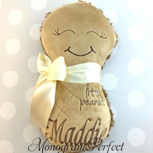 Puede incluir: Un juguete de peluche color canela con forma de cacahuete, con ojos cerrados y una boca sonriente bordados. El juguete tiene una cinta de sat&eacute;n color crema atada en el medio, con el texto bordado "little peanut" y el nombre "Maddie".
