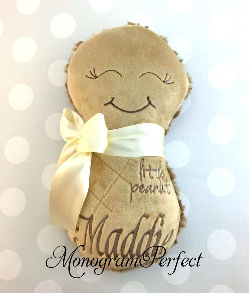 Puede incluir: Un juguete de peluche color canela con forma de cacahuete, con ojos cerrados y una boca sonriente bordados. El juguete tiene una cinta de sat&eacute;n color crema atada en el medio, con el texto bordado "little peanut" y el nombre "Maddie".