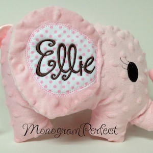 Puede incluir: Elefante de peluche rosa con un nombre bordado en marrón "Ellie" en un círculo de lunares blancos y rosas. El elefante tiene una cola de satén rosa.