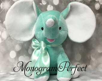 May include: A plush elephant toy in mint green with large white inner ears, a white tip on its trunk, and white feet. A matching mint green satin ribbon is tied around its neck. The text Monogram Perfect is printed across the lower portion.