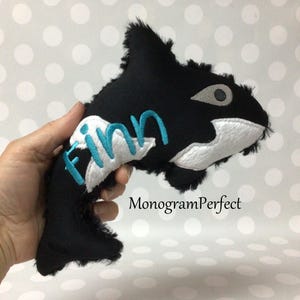 May include: A black and white orca whale soft toy with the name "Finn" embroidered in blue. The toy has a furry texture and a grey eye. The text "MonogramPerfect" is printed below the toy.