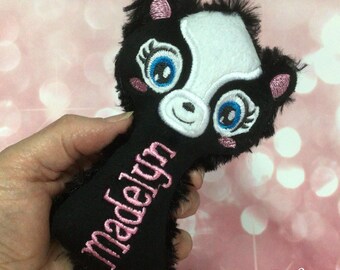May include: A black and white plush skunk toy with blue eyes and pink accents. The name "Madelyn" is embroidered in pink on the skunk's body. The toy has a fluffy texture and is held in a hand.