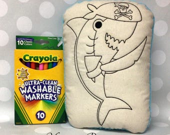 May include: A plush shark toy with a pirate hat and a crayon box. The shark is outlined in black on a cream-colored background with a light blue border. The crayon box contains 10 classic colors.