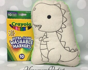 May include: A beige dinosaur-shaped pillow with black outline details and a green border, next to a box of Crayola Ultra-Clean Washable Markers. The box contains 10 classic colors.