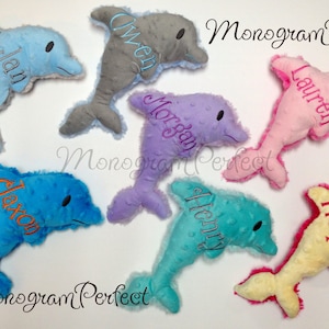 May include: A selection of soft dolphin toys in shades of blue, grey, purple, pink, turquoise, and yellow. Each dolphin features an embroidered name in a contrasting colour. The text "Monogram Perfect" is also visible.