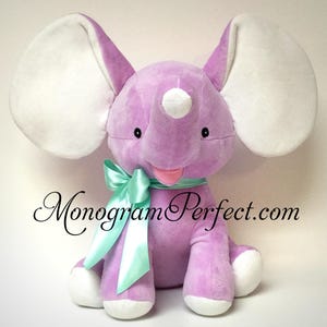Può includere: Un peluche a forma di elefante di colore viola chiaro con grandi orecchie interne bianche, piedi bianchi e una punta bianca sulla proboscide. L'elefante presenta un nastro di raso verde menta annodato al collo e il testo MonogramPerfect.com.
