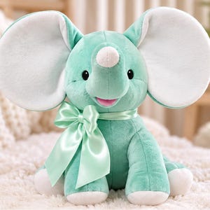 May include: A plush elephant toy in a soft mint green colour with large, rounded ears featuring white inner panels. The stuffed animal has a small white horn on its forehead, black embroidered eyes, a pink mouth, and a satin mint green bow.