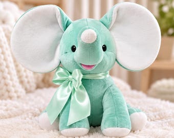 May include: A plush elephant toy in a soft mint green color with large, rounded ears featuring white inner panels. The stuffed animal has a small white horn on its forehead, black embroidered eyes, a pink mouth, and a satin mint green bow.