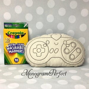 Puede incluir: Un cojín hecho a mano de color crema con forma de mando de videojuegos, con detalles cosidos en negro. Junto a él, una caja de rotuladores lavables Crayola Ultra-Clean, 10 colores clásicos. El texto "Monogram Perfect" está en la parte inferior.