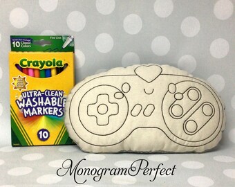 May include: A cream-colored, handmade pillow in the shape of a video game controller, with black stitched details. Next to it is a box of Crayola Ultra-Clean Washable Markers, 10 classic colors. The text "Monogram Perfect" is at the bottom.