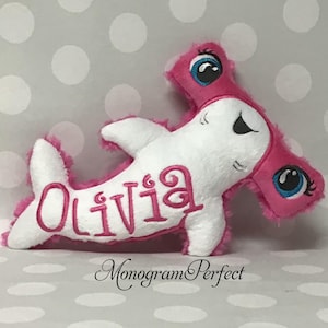 May include: A soft toy hammerhead shark in white and pink. The shark features large blue eyes, a smiling face, and the name "Olivia" embroidered in pink. The toy has a fluffy texture and pink trim.
