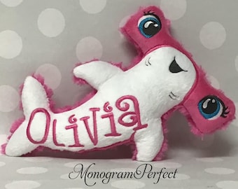May include: A plush hammerhead shark toy in white and pink. The shark has large blue eyes, a smiling face, and the name "Olivia" embroidered in pink. The toy is soft and fluffy, with a pink trim.